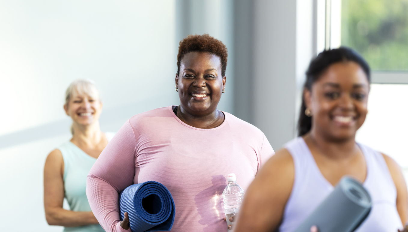 three women smile after yoga class holding their mats and water