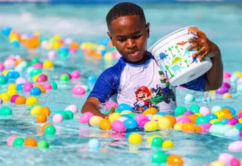 Boy in wet swimming clothes holding a basket in the pool surrounded by colorful plastic eggs
